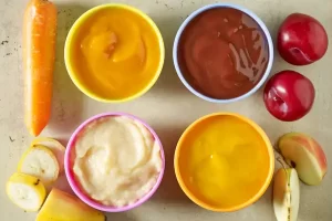 O momento de iniciar a alimentação complementar do bebê traz consigo uma mistura de emoções: alegria por alcançar mais um marco no desenvolvimento, ansiedade por fazer escolhas alimentares adequadas e, por vezes, preocupação quando notamos que nosso pequeno apresenta desconfortos digestivos. Para famílias lidando com bebês que demonstram sensibilidade intestinal leve, a jornada alimentar precisa de um olhar especial, particularmente quando se trata da introdução de fibras – componentes essenciais para o desenvolvimento de um sistema digestivo equilibrado. A relação entre a alimentação e o conforto digestivo de bebês é delicada e personalizada. Cada pequeno corpo responde de maneira única aos alimentos, e o ritmo de adaptação às fibras pode variar consideravelmente. Este cuidado particularizado não significa privar o bebê de nutrientes importantes, mas sim apresentá-los de forma gradual e estratégica, respeitando o tempo que o organismo necessita para se adaptar. Entendendo a Sensibilidade Intestinal em Bebês A sensibilidade intestinal leve em bebês pode se manifestar de diversas formas: gases em excesso, cólicas ocasionais, alterações no padrão das fezes ou desconforto após determinadas refeições. Diferente de condições como alergias alimentares ou intolerâncias diagnosticadas, esta sensibilidade representa uma reação temporária do sistema digestivo ainda em desenvolvimento. O trato gastrointestinal do bebê passa por um período de amadurecimento intenso durante o primeiro ano de vida. A microbiota intestinal – conjunto de microrganismos que habitam nosso intestino – está em formação, e o contato com novos alimentos representa um estímulo importante para este processo. As fibras, em particular, contribuem significativamente para a diversificação desta microbiota, mas quando introduzidas abruptamente ou em quantidade inadequada, podem causar desconforto. Sinais de Sensibilidade Intestinal que Merecem Atenção Alguns sinais podem indicar que o bebê apresenta sensibilidade intestinal leve e que se beneficiaria de uma introdução gradual de fibras: Distensão abdominal visível após as refeições Inquietação durante ou logo após alimentação Gases frequentes acompanhados de desconforto Fezes muito duras ou muito moles Recusa alimentar associada a determinados alimentos Estudos publicados no Journal of Pediatric Gastroenterology and Nutrition indicam que aproximadamente 25% dos bebês apresentam algum grau de sensibilidade digestiva durante a introdução alimentar, sendo que a maioria dos casos se resolve com ajustes na dieta e com o próprio amadurecimento natural do sistema digestivo. O Papel das Fibras na Alimentação Infantil As fibras são componentes não digeríveis dos alimentos vegetais que desempenham funções essenciais no organismo. Conforme pesquisas da Academia Americana de Pediatria, uma alimentação adequada em fibras desde a primeira infância está associada a benefícios de curto e longo prazo, como: Regulação do trânsito intestinal Prevenção de constipação Alimentação da microbiota intestinal benéfica Desenvolvimento de preferências por alimentos naturais Estabelecimento de hábitos alimentares associados ao bem-estar duradouro na vida adulta Existem dois tipos principais de fibras, cada uma com seus benefícios: Fibras solúveis: Absorvem água, formam géis e tendem a atrasar o esvaziamento gástrico. Estão presentes em frutas como maçã, pera, banana, aveia, cevada e leguminosas. Para bebês com sensibilidade intestinal, estas costumam ser melhor toleradas inicialmente. Fibras insolúveis: Não absorvem água significativamente e ajudam a dar volume às fezes. Encontradas principalmente em vegetais folhosos, cereais integrais e cascas de frutas. Devem ser introduzidas mais gradualmente em bebês sensíveis. Estratégias para Introdução Progressiva de Fibras A abordagem progressiva na introdução de fibras baseia-se na compreensão de que o sistema digestivo do bebê precisa de tempo para se adaptar. Pesquisas conduzidas pelo Departamento de Nutrição Pediátrica da Universidade de Leeds demonstram que a introdução gradual de alimentos com fibras pode reduzir em até 60% os desconfortos digestivos em bebês com sensibilidade intestinal leve. Fase 1: O Início Suave (6-7 meses) Nesta fase inicial, o foco está em alimentos com baixo teor de fibras ou com fibras muito bem processadas: Vegetais de baixo teor de fibras: Abóbora cozida e peneirada Batata doce bem cozida e amassada Abobrinha sem casca e sem sementes, cozida até ficar muito macia Cenoura bem cozida e processada finamente Frutas de fácil digestão: Banana madura amassada Pera madura, sem casca, cozida e amassada Maçã ralada e cozida até ficar bem macia Mamão papaia maduro amassado Exemplo de progressão: Comece com purês de um único ingrediente, como abóbora pura Após 2-3 dias sem reações, experimente outro ingrediente isoladamente Depois de testar vários alimentos individualmente, inicie combinações simples Receita exemplar: Purê de Abóbora Aveludado 1 xícara de abóbora japonesa descascada e cortada em cubos pequenos Água filtrada suficiente para cobrir Preparo: Cozinhe a abóbora em pouca água até ficar bem macia (cerca de 15 minutos). Processo no liquidificador ou amasse bem com um garfo até obter um purê completamente liso. Para os primeiros contatos com papinhas, a textura extremamente homogênea facilita a digestão. Fase 2: Ampliando o Repertório com Cautela (7-9 meses) Nesta fase, começamos a aumentar gradualmente o teor de fibras e a complexidade das preparações: Vegetais com teor moderado de fibras: Brócolis (apenas os floretes bem cozidos) Couve-flor (floretes cozidos até ficarem muito macios) Beterraba cozida Abóbora com menos processamento (textura um pouco mais grosseira) Frutas com mais fibras: Maçã com casca cozida (bem macia e depois processada) Ameixa sem casca e cozida Pêssego maduro sem casca e cozido Cereais e grãos leves: Aveia em flocos finos cozida até formar um mingau cremoso Arroz bem cozido e amassado Milhinho bem cozido Receita exemplar: Mingau de Aveia com Pera e Canela 2 colheres de sopa de aveia em flocos finos 1/2 pera madura, sem casca e sem sementes 1 pitada de canela em pó 150ml de água filtrada Preparo: Cozinhe a aveia na água por aproximadamente 5 minutos, mexendo sempre. Adicione a pera cortada em pedaços pequenos e cozinhe por mais 3 minutos. Quando estiver cremoso e a pera estiver macia, deixe esfriar um pouco e adicione uma pitada de canela. Processe conforme a textura adequada para a idade do bebê. Este mingau oferece uma combinação de fibras solúveis da aveia e da pera, com a canela que, além de saborizar naturalmente, tem propriedades que auxiliam na digestão. A textura homogênea e a cocção adequada facilitam o processo digestivo. Fase 3: Diversificação Consciente (9-12 meses) Nesta fase, podemos aumentar gradualmente o teor de fibras e introduzir texturas mais complexas, sempre observando a tolerância individual do bebê: Leguminosas bem cozidas: Lentilha vermelha (dhal) cozida até desmanchar Grão-de-bico bem cozido e amassado Feijão carioca sem casca e bem cozido Cereais com maior teor de fibras: Quinoa bem cozida Aveia em flocos regulares Arroz integral bem cozido e amassado Vegetais mais fibrosos (bem cozidos): Espinafre refogado e picado finamente Acelga bem cozida Ervilhas amassadas Receita exemplar: Risotinho de Legumes com Lentilha 2 colheres de sopa de arroz 1 colher de sopa de lentilha vermelha 1/4 de cenoura pequena picada 1/4 de abobrinha pequena sem casca e sem sementes, picada 1 colher de chá de azeite de oliva extra virgem 1/2 xícara de água filtrada ou caldo de legumes caseiro Preparo: Lave bem o arroz e a lentilha. Em uma panela pequena, coloque um pouco de água e adicione o arroz, a lentilha e os legumes picados. Cozinhe em fogo baixo até que todos os ingredientes estejam bem macios (aproximadamente 20-25 minutos), adicionando mais água se necessário. Quando tudo estiver bem cozido, amasse com um garfo ou processe brevemente, mantendo um pouco de textura se o bebê já estiver aceitando alimentos menos homogêneos. Finalize com o azeite. Esta receita combina diferentes fontes de fibras (lentilha, arroz, legumes) em uma preparação que, devido ao cozimento adequado, torna-se mais digerível. A lentilha vermelha é particularmente interessante para bebês sensíveis, pois se desmancha facilmente durante o cozimento. Técnicas de Preparo que Favorecem a Digestibilidade O modo de preparo dos alimentos pode influenciar significativamente sua tolerância digestiva. Pesquisas publicadas no European Journal of Clinical Nutrition demonstram que técnicas culinárias adequadas podem reduzir fatores antinutricionais e aumentar a biodisponibilidade de nutrientes, além de melhorar a digestibilidade. Algumas técnicas particularmente úteis incluem: Cocção Prolongada e em Baixa Temperatura Para leguminosas e vegetais fibrosos, a cocção prolongada em fogo baixo ajuda a quebrar as fibras mais resistentes, tornando-as mais digeríveis. No caso do feijão, por exemplo, o remolho prévio seguido de descarte da água e cocção prolongada pode reduzir significativamente componentes que causam flatulência. Uso Estratégico de Especiarias Digestivas Certas especiarias tradicionalmente utilizadas na culinária mundial têm propriedades que auxiliam na digestão: Cominho: auxilia na digestão de leguminosas Erva-doce: combate gases e cólicas Gengibre (em pequenas quantidades): estimula enzimas digestivas Canela: auxilia no controle glicêmico e tem ação anti-inflamatória Fermentação Natural Processos fermentativos controlados podem pré-digerir parcialmente componentes complexos dos alimentos. Por exemplo: Iogurte natural caseiro (após os 6 meses, com orientação pediátrica) Mingau de aveia deixado de molho na água (overnight oats adaptado para bebês) Receita exemplar: Purê de Maçã Fermentado 2 maçãs maduras, de preferência orgânicas 1 colher de chá de suco de limão Preparo: Descasque e corte as maçãs em pedaços pequenos. Coloque em uma panela com um pouco de água e cozinhe até ficarem macias. Amasse bem e deixe esfriar até ficar morno. Adicione o suco de limão, transfira para um pote de vidro esterilizado, tampado, mas não completamente fechado, e deixe em temperatura ambiente por 8-12 horas. Depois refrigere. Este processo permite uma fermentação leve que pode auxiliar na digestibilidade. Acompanhando as Reações do Bebê A observação atenta das reações do bebê após a introdução de novos alimentos é fundamental para personalizar o ritmo de introdução das fibras. Um diário alimentar pode ser uma ferramenta valiosa neste processo. Nele, pode-se registrar: Alimentos oferecidos e quantidades aproximadas Aceitação e rejeição Horário das refeições Comportamento após a alimentação (sonolência, irritabilidade, etc.) Padrão de evacuação (frequência, consistência, cor) Sinais de desconforto abdominal Analisando estes registros ao longo do tempo, torna-se possível identificar padrões e fazer ajustes personalizados. Cardápio Semanal Progressivo Um planejamento semanal facilita a organização e garante a variedade nutricional necessária. Aqui está um exemplo de cardápio progressivo para cada fase: Cardápio Fase 1 (6-7 meses) Segunda: Manhã: Purê de banana madura Tarde: Purê de batata doce Terça: Manhã: Purê de pera cozida Tarde: Purê de abóbora Quarta: Manhã: Purê de maçã cozida Tarde: Purê de cenoura Quinta: Manhã: Purê de banana com um pouco de aveia bem cozida Tarde: Purê de batata doce com abobrinha Sexta: Manhã: Purê de mamão Tarde: Mistura de abóbora com cenoura Sábado: Manhã: Purê de pera com um pouco de aveia Tarde: Purê de batata inglesa Domingo: Manhã: Mistura de frutas já testadas (banana e maçã, por exemplo) Tarde: Mistura de vegetais já testados (batata doce e cenoura) Cardápio Fase 2 (7-9 meses) Segunda: Manhã: Mingau de aveia com banana Almoço: Purê de abóbora com frango desfiado Lanche: Purê de maçã com canela Terça: Manhã: Papa de arroz bem cozido com cenoura Almoço: Purê de batata com brócolis bem cozido Lanche: Purê de pera com ameixa Quarta: Manhã: Mingau de milhinho Almoço: Purê de abobrinha com tilápia Lanche: Banana amassada com aveia em flocos finos Quinta: Manhã: Aveia com maçã ralada cozida Almoço: Purê de batata doce com espinafre e gema de ovo cozida Lanche: Purê de mamão com pera Sexta: Manhã: Papa de arroz com abóbora Almoço: Purê de cenoura com couve-flor e frango Lanche: Purê de pêssego Sábado: Manhã: Mingau de aveia com canela Almoço: Purê de batata com beterraba e carne moída Lanche: Maçã ralada cozida com pera Domingo: Manhã: Papa de arroz com cenoura Almoço: Purê misto de legumes com proteína já testada Lanche: Mistura de frutas já testadas Cardápio Fase 3 (9-12 meses) Segunda: Manhã: Mingau de aveia com frutas picadas Almoço: Risoto de frango com cenoura e abobrinha Lanche: Banana amassada com aveia Jantar: Sopa de legumes com macarrão infantil Terça: Manhã: Papa de quinoa com maçã Almoço: Purê rústico de batata com espinafre e peixe Lanche: Pera em pedaços pequenos com canela Jantar: Sopa de abóbora com lentilha vermelha Quarta: Manhã: Mingau de aveia com banana e chia hidratada Almoço: Arroz bem cozido com lentilha e legumes Lanche: Mamão em pedaços pequenos Jantar: Creme de ervilha com batata Quinta: Manhã: Papa de tapioca com frutas Almoço: Purê de batata doce com brócolis e frango Lanche: Iogurte natural com frutas amassadas (se já liberado pelo pediatra) Jantar: Sopa de legumes com arroz Sexta: Manhã: Mingau de aveia com pêssego Almoço: Arroz integral bem cozido com feijão sem casca e carne desfiada Lanche: Maçã ralada com aveia Jantar: Creme de abóbora com cenoura Sábado: Manhã: Papa de quinoa com frutas Almoço: Polenta mole com molho de tomate natural e frango Lanche: Banana em rodelas com aveia polvilhada Jantar: Sopa de legumes variados Domingo: Manhã: Mingau de aveia com mix de frutas Almoço: Arroz, feijão bem cozido, carne e legumes (versão família adaptada) Lanche: Frutas picadas variadas Jantar: Creme de legumes com macarrão infantil Dicas para Situações Especiais Quando o Bebê Recusa Alimentos com Fibras A recusa alimentar pode ter diversas causas, desde questões sensoriais até experiências prévias de desconforto. Algumas estratégias incluem: Mascarar alimentos mais fibrosos com sabores já conhecidos e aceitos pelo bebê Variar a temperatura dos alimentos (alguns bebês preferem alimentos mais mornos, outros à temperatura ambiente) Oferecer em momentos de maior fome Transformar o momento da refeição em uma experiência positiva, sem pressão Lidando com Constipação A constipação pode ocorrer durante a alimentação complementar, especialmente durante as transições entre fases: Aumentar temporariamente a oferta de alimentos com efeito laxativo natural, como ameixa, mamão papaia e abacate Garantir hidratação adequada Massagem abdominal no sentido horário Exercícios de movimentação das pernas tipo "bicicleta" Um estudo publicado no Journal of Family Practice demonstrou que simples ajustes na dieta resolveram casos de constipação em 80% dos bebês estudados, sem necessidade de medicamentos. Quando Procurar Ajuda Profissional Algumas situações merecem avaliação por profissionais de saúde: Constipação persistente (mais de uma semana) Diarreia (fezes líquidas em maior frequência que o habitual) Presença de muco ou sangue nas fezes Dor abdominal intensa com choro inconsolável Vômitos persistentes Perda de peso ou parada no ganho de peso Reações cutâneas como urticária após determinados alimentos Mitos e Verdades sobre Fibras na Alimentação de Bebês Mito 1: Bebês não precisam de fibras nos primeiros meses da alimentação complementar. Realidade: A introdução gradual de fibras é importante desde o início da alimentação complementar, pois contribui para o desenvolvimento saudável do sistema digestivo e da microbiota intestinal. Mito 2: Alimentos com fibras sempre causam gases e desconforto. Realidade: Quando introduzidos gradualmente e preparados adequadamente, os alimentos fibrosos são geralmente bem tolerados. O desconforto costuma estar mais associado à quantidade e à forma de preparo do que ao alimento em si. Mito 3: Bebês com sensibilidade intestinal devem evitar completamente alimentos integrais. Realidade: Não é necessária a exclusão completa, mas sim uma introdução mais cautelosa e progressiva, com técnicas de preparo que favoreçam a digestibilidade. Mito 4: Só é possível oferecer fibras quando o bebê já tem dentes. Realidade: As fibras podem ser oferecidas em texturas adequadas à fase do bebê, mesmo antes do surgimento dos primeiros dentes, desde que devidamente processadas. A Jornada Contínua da Alimentação Infantil A alimentação do bebê é uma jornada dinâmica que se transforma constantemente. Ao adotar uma abordagem progressiva na introdução de fibras, estamos não apenas minimizando possíveis desconfortos digestivos, mas também construindo as bases para uma relação saudável com a comida que persistirá ao longo da vida. Pesquisas conduzidas no King's College London demonstram que as experiências alimentares nos primeiros 1000 dias de vida (da concepção aos 2 anos) têm impacto duradouro no equilíbrio metabólico, nas preferências alimentares e até mesmo no comportamento alimentar na vida adulta. Ao respeitar o ritmo individual de cada bebê e ao mesmo tempo promover gradualmente a diversidade alimentar, oferecemos um presente valioso: o desenvolvimento de um sistema digestivo resiliente e uma relação positiva com alimentos naturais e nutritivos. Lembre-se sempre: cada bebê é único, com seu próprio tempo e suas particularidades fisiológicas. A paciência, a observação atenta e a flexibilidade para ajustar o plano alimentar são ingredientes tão essenciais quanto os próprios alimentos que oferecemos aos nossos pequenos. A introdução progressiva de fibras não é apenas uma estratégia para minimizar desconfortos; é uma forma de respeito ao desenvolvimento individual e ao processo de descoberta alimentar de cada criança.